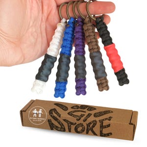 Könnte beinhalten: Fünf Paracord-Schlüsselanhänger mit bronzefarbenen Ringen, jeweils mit einem geflochtenen Kordelabschnitt in Weiß, Blau, Lila, Braun oder Schwarz und einem einfarbigen Gummigriff. Darunter befindet sich ein brauner Karton mit der Aufschrift BJJ FAMILY BUSINESS.