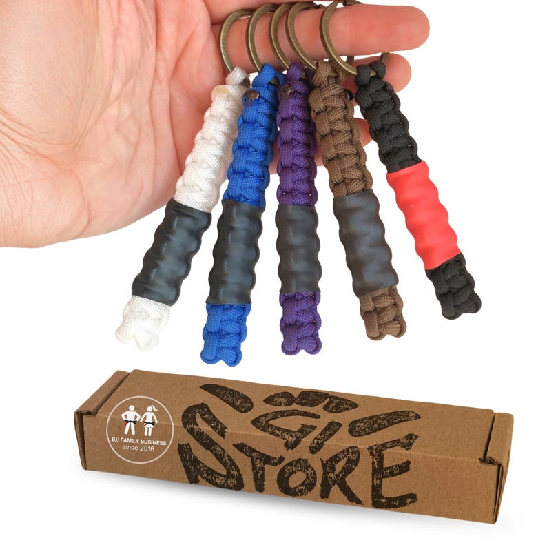 May include: Five paracord keychains with bronze-toned rings, each featuring a woven cord section in white, blue, purple, brown, or black, paired with a solid-colored rubber grip. Below sits a brown cardboard box labeled BJJ FAMILY BUSINESS.