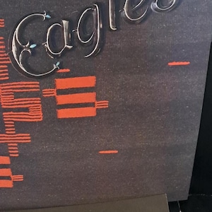 May include: Two Eagles album covers are displayed. The top cover has a dark background with an orange geometric pattern and the band's name in a silver, stylized font. The bottom cover features a band photo and the band's name.