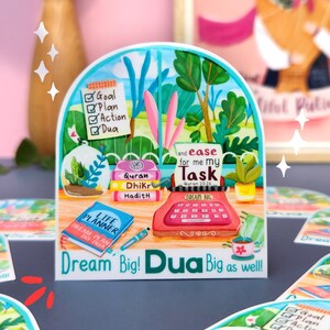 May include: A colorful sticker with a typewriter, books, and plants. The sticker has the text "Dream Big! Dua Big as well!" and "and ease for me my Task Quran 20:26".