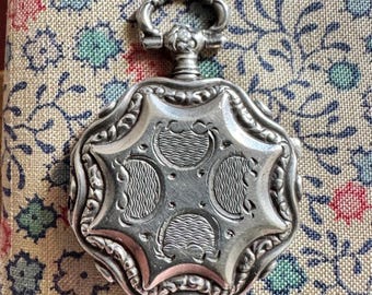 May include: A silver pocket watch with an ornate, star-shaped design. The watch features a detailed, engraved face with a circular pattern. The watch is set against a floral patterned fabric with red, blue, and green flowers.