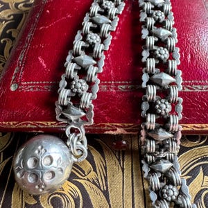 May include: A silver-toned metal chain bracelet featuring alternating rectangular links with embossed floral and diamond-shaped patterns. The bracelet terminates in a large, spherical silver-toned charm engraved with a symmetrical four-petal floral design.