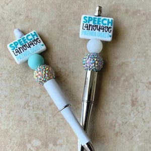May include: Two pens featuring white barrels and silver accents. Each pen is topped with a rectangular white bead printed with the text SPEECH LANGUAGE PATHOLOGIST in blue and black, a solid light blue bead, and a sparkling rhinestone bead.