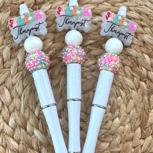 May include: Three white glitter pens featuring a topper shaped like a speech bubble with the text Speech Therapist in colourful letters. Each pen includes a white bead and a pink and yellow rhinestone-encrusted bead above the white barrel.