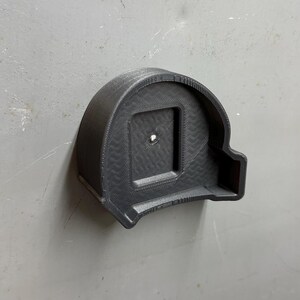Pu&ograve; includere: Un supporto nero per metro a nastro stampato in 3D. Il supporto &egrave; montato su una superficie grigia e presenta un'apertura quadrata al centro. Il design include un motivo strutturato e un piccolo foro per il montaggio. Il supporto &egrave; progettato per contenere saldamente un metro a nastro.