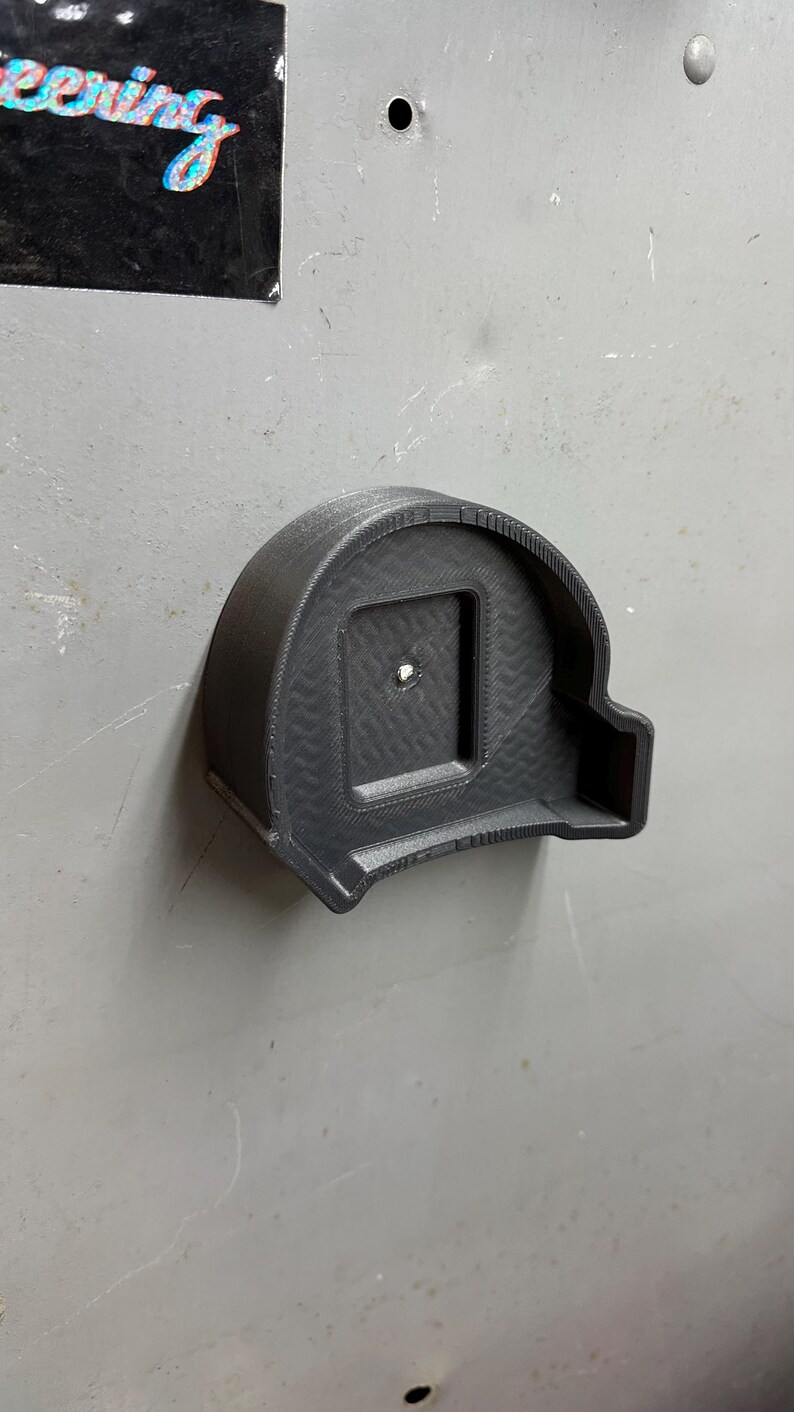 Pu&ograve; includere: Un supporto nero per metro a nastro stampato in 3D. Il supporto &egrave; montato su una superficie grigia e presenta un'apertura quadrata al centro. Il design include un motivo strutturato e un piccolo foro per il montaggio. Il supporto &egrave; progettato per contenere saldamente un metro a nastro.