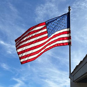 Puede incluir: Una bandera estadounidense roja, blanca y azul ondeando en el viento contra un cielo azul brillante con nubes blancas.