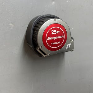 Pu&ograve; includere: Un metro a nastro Snap-on nero e argento da 7,62 metri con un centro rosso. Il metro a nastro ha il testo "25FT Snap-on TPMA25" stampato sul centro rosso. Il metro a nastro &egrave; su una superficie grigia.