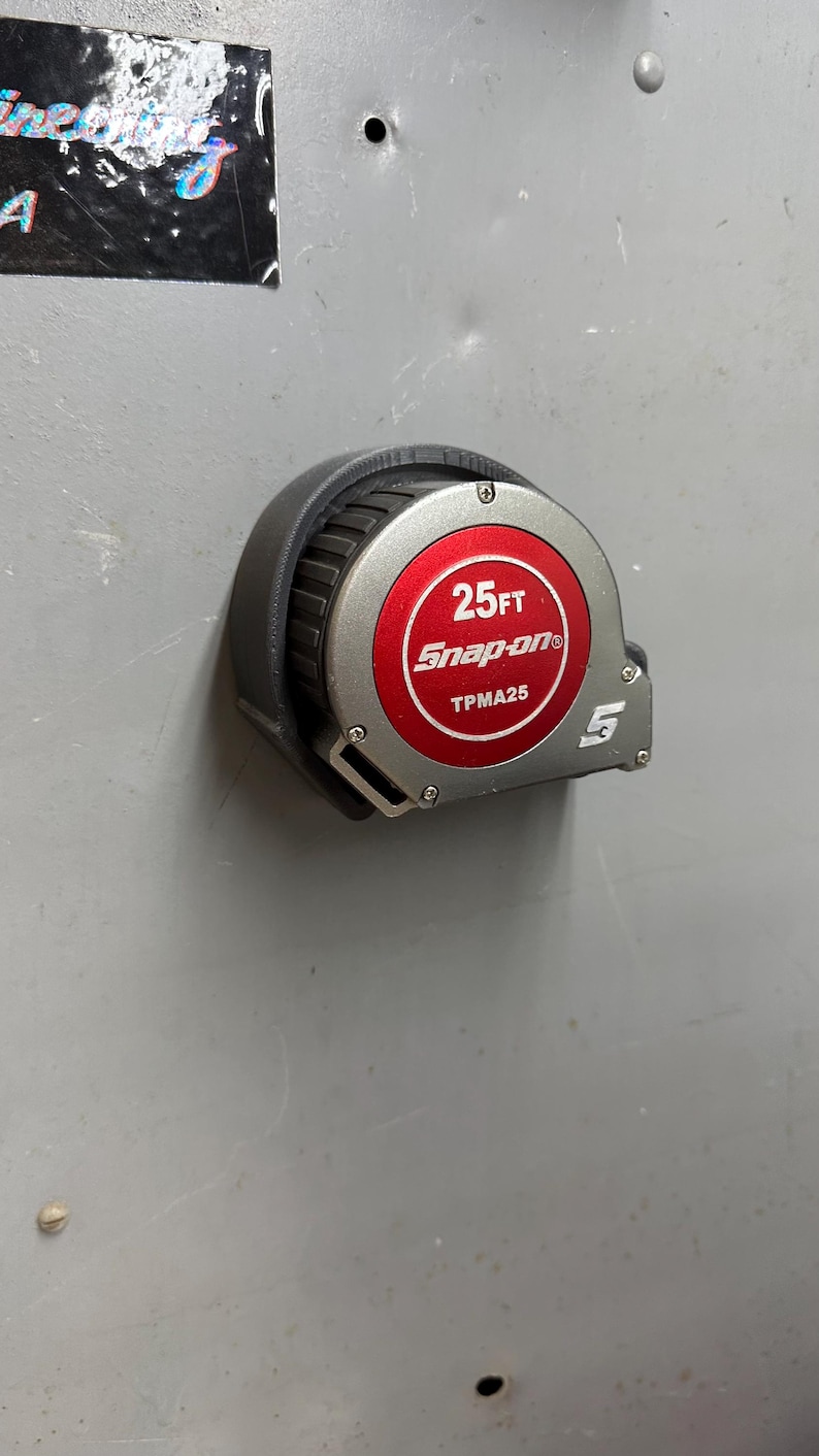 Pu&ograve; includere: Un metro a nastro Snap-on nero e argento da 7,62 metri con un centro rosso. Il metro a nastro ha il testo "25FT Snap-on TPMA25" stampato sul centro rosso. Il metro a nastro &egrave; su una superficie grigia.