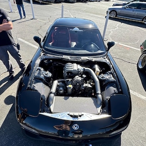 Pode incluir: Um Mazda RX-7 preto com o capô aberto, revelando o motor. O carro está estacionado em um estacionamento com outros carros ao fundo.