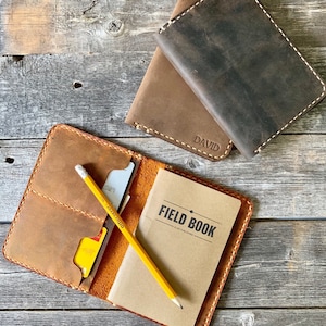 May include: A brown leather notebook cover with a yellow pencil and a small notebook that says "FIELD BOOK" inside. The cover has a pocket for cards and a stitched design.