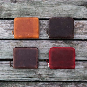 May include: Four leather zippered wallets in different colors: brown, dark brown, red, and black. The wallets are arranged in two rows of two on a wooden surface.