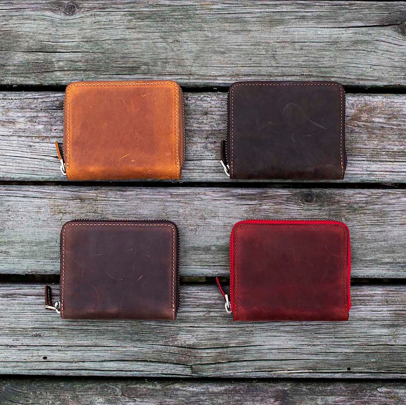 May include: Four leather zippered wallets in different colors: brown, dark brown, red, and black. The wallets are arranged in two rows of two on a wooden surface.