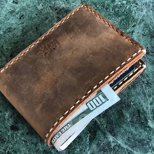 May include: A brown leather wallet with white stitching and a bill sticking out. The wallet has the text "Leather" embossed on the front.