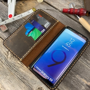 May include: A brown leather phone case with stitching around the edges. The case has a slot for credit cards and a flap that covers the phone. A Samsung Galaxy S9 phone is inside the case. The case is on a wooden surface.