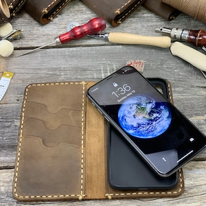 May include: Open brown leather phone case with card slots, holding a black smartphone displaying a world map wallpaper. The case is stitched with light-colored thread. Other leather wallets and tools are in the background.