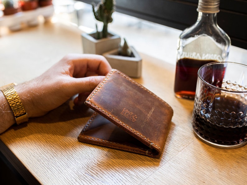 May include: A brown leather wallet with the text "Lone & Rebel" embossed on the front. The wallet is folded in half and is being held by a hand.