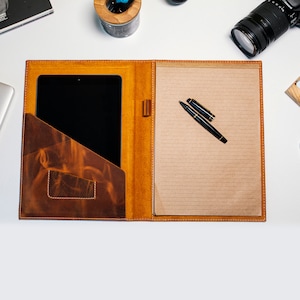 May include: A brown leather portfolio with a flap closure and a pocket for a tablet or notepad. The portfolio has a lined notepad and two black pens inside.