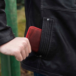 May include: A red leather wallet with a zipper is being placed in the pocket of a black leather jacket.