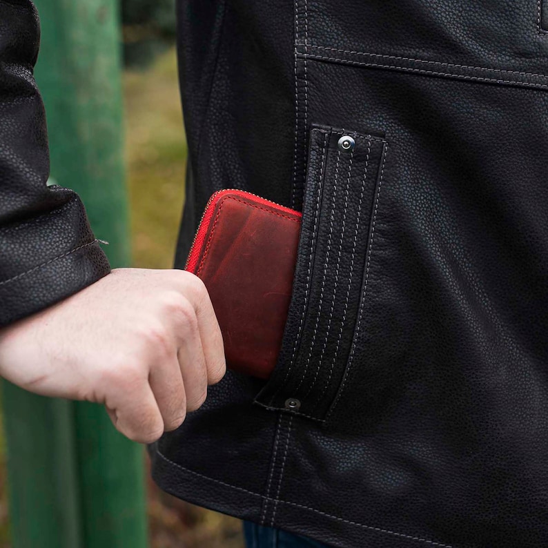 May include: A red leather wallet with a zipper is being placed in the pocket of a black leather jacket.
