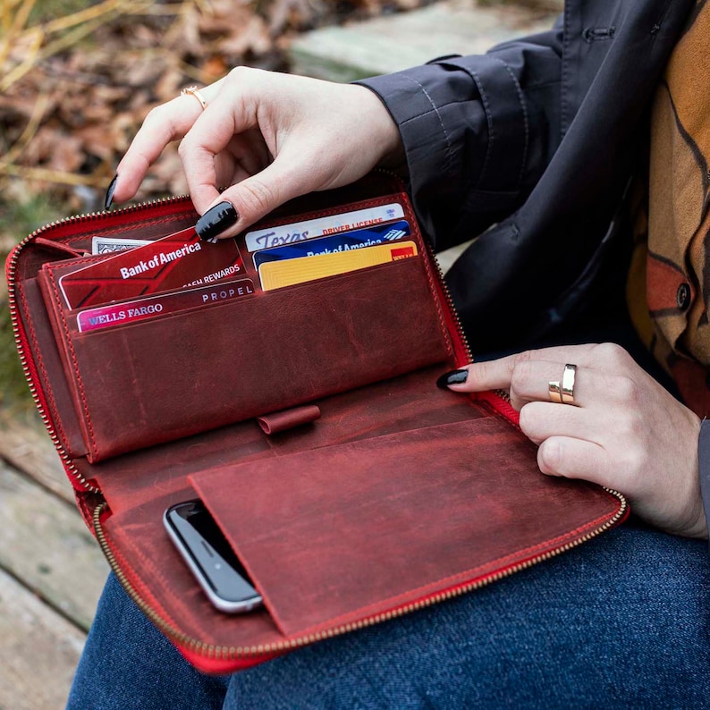 May include: A red leather zippered wallet with multiple card slots and a phone pocket. The wallet is open and has several credit cards inside, including a Bank of America card, a Wells Fargo card, and a Texas driver's license.