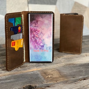 May include: A brown leather phone case with a built-in wallet. The case is open and a smartphone with a screen displaying "Note 10 Pro" is visible. The case has slots for credit cards and cash.