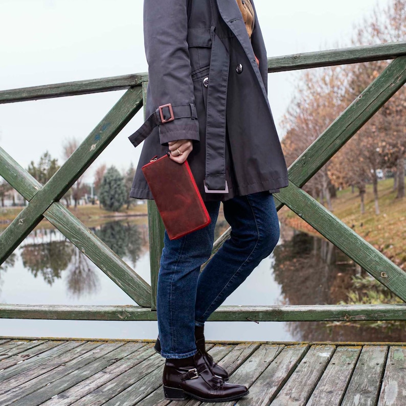 May include: A woman wearing a gray trench coat and blue jeans holds a red leather clutch purse.