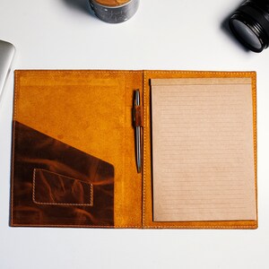 May include: A brown leather folder with a notepad and pen. The folder has a pocket on the left side and a flap that closes with a snap.