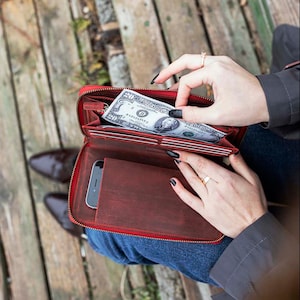 May include: A red leather wallet with a zipper closure. The wallet is open and a dollar bill is visible inside. The wallet is sitting on a wooden surface.