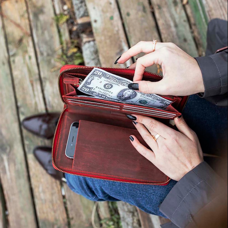 May include: A red leather wallet with a zipper closure. The wallet is open and a dollar bill is visible inside. The wallet is sitting on a wooden surface.