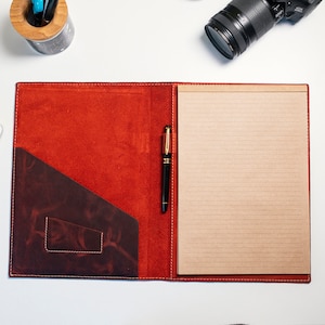 May include: A red leather folder with a brown leather pocket on the front. The folder is open to reveal a lined sheet of paper and a black pen.