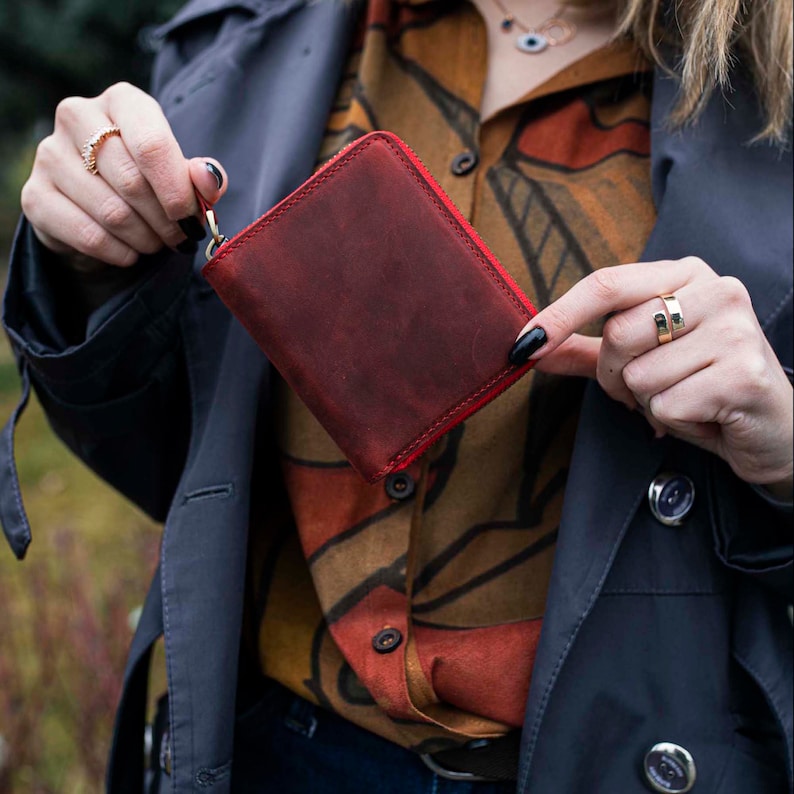 May include: A red leather wallet with a zipper closure. The wallet is being held by a person's hand.