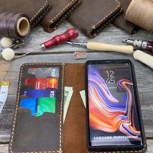 May include: A brown leather phone case with a stitched edge and a slot for credit cards. The case is open and a smartphone with a colorful screen is visible. The phone screen displays the time 12:45 and the text "SAMSUNG Galaxy Note9".