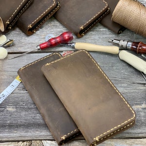 May include: Handmade leather wallets in various shades of brown, with visible stitching in a contrasting cream color. The wallets are arranged with leatherworking tools, including an awl, thread, and a measuring tape, suggesting a craft theme.