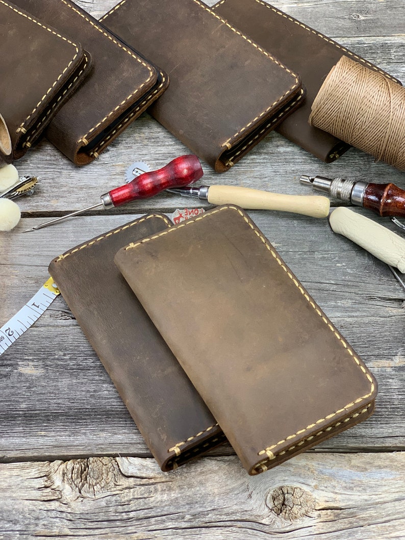 May include: Handmade leather wallets in various shades of brown, with visible stitching in a contrasting cream color. The wallets are arranged with leatherworking tools, including an awl, thread, and a measuring tape, suggesting a craft theme.