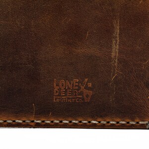 May include: Close-up of a brown leather wallet with a stitched edge. The wallet has a logo embossed on the front that reads "Lone Deer Leather Co."