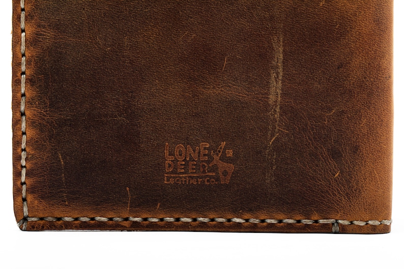 May include: Close-up of a brown leather wallet with a stitched edge. The wallet has a logo embossed on the front that reads "Lone Deer Leather Co."
