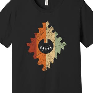 Puede incluir: Camiseta negra con un diseño geométrico desgastado en naranja, beige y verde. El diseño incluye un círculo central con pequeñas figuras blancas. La camiseta tiene cuello redondo y mangas cortas.