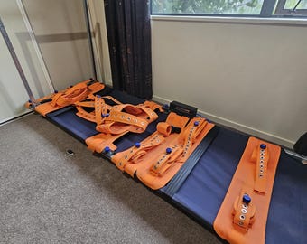 May include: A blue and orange medical stretcher with multiple orange straps and buckles. The stretcher is on a gray carpeted floor, near a window and a metal frame.