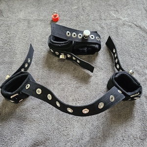 Puede incluir: Tres ataduras de muñeca de tela negra con ojales metálicos y una hebilla plateada. Las ataduras están hechas de una tela suave y acolchada y tienen un lazo para sujetar a otro objeto.