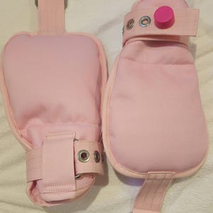 Könnte beinhalten: Zwei hellrosa gepolsterte Fesseln mit verstellbaren Riemen und Metallbeschlägen. Eine Fessel hat einen rosa Knopfverschluss. Sie sind für die Sicherung von Handgelenken oder Knöcheln konzipiert und aus einem weichen, strapazierfähigen Stoff gefertigt.