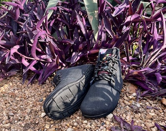 May include: A pair of black canvas boots with black and white patterned laces. The boots are placed on a bed of small pebbles, with purple and green plants in the background.