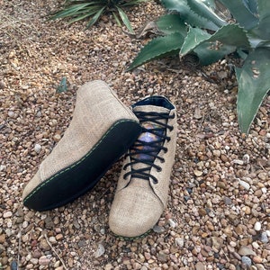 Puede incluir: Par de botas de yute color beige con suelas negras y cordones negros. Las botas tienen un diseño de caña alta con un forro estampado colorido. Las suelas están cosidas con hilo verde. Una bota está tumbada de lado.