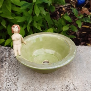 Może przedstawiać: Mała zielona ceramiczna miska z otworem odpływowym na dnie. Mała ceramiczna figurka kobiety z czerwonymi włosami siedzi na krawędzi miski.