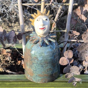 May include: A handcrafted ceramic sculpture of a whimsical figure with a sun-like crown and a smiling face. The figure has a teal-colored body with spiral designs and a ruffled collar. The sculpture is set against a natural background with branches and foliage.