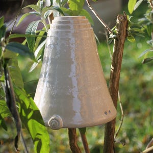 Pode incluir: Um sino de cerâmica branco com uma superfície texturizada pendurado em um galho em um jardim. O sino tem um topo arredondado e uma base larga.
