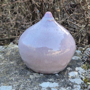 Könnte beinhalten: Helllila Keramik-Dekorationsobjekt mit rundem Körper und spitzer Spitze. Das Objekt hat eine glänzende Oberfläche und steht auf einer grauen Steinoberfläche. Der Hintergrund zeigt kahle Äste.