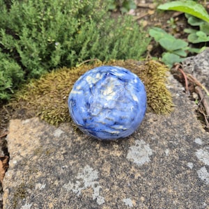 Può includere: Un oggetto sferico in ceramica con finitura lucida, caratterizzato da un motivo marmorizzato nei toni del blu scuro, azzurro e bianco. La superficie presenta un aspetto irregolare e strutturato con sottili rientranze e rilievi.