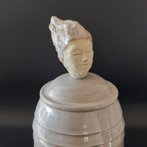 Può includere: Un vaso di ceramica con un coperchio a forma di testa umana. Il vaso è di colore grigio chiaro e la testa è di colore marrone chiaro. La testa ha gli occhi chiusi e un'espressione serena.