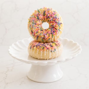May include: Two stacked ring-shaped pastries with white icing and colorful sprinkles on a white scalloped pedestal stand. The top pastry is heavily covered in pink, yellow, blue, and purple sprinkles, while the bottom pastry has fewer.